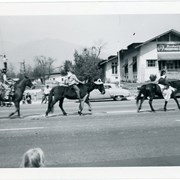 Three youngsters riding their horses along parade route for March 1949 Peach Blossom Festival Parade.  They are passing approximately 27 West Huntington Drive, as the sign for Barbour's Preserving Co. is visible above the canopy.