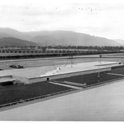 View NE across enormous warehouse nearing completion on infield area of track at Santa Anita Assembly Center.  Houses seen across top of warehouse would be in what was called Santa Anita Gardens area.  Santa Anita Dam can be faintly seen in prominent fold of the mountains. This photo, together with photo #653, forms a panorama.