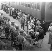 Fourteen soldiers stand guard alongside Pacific Electric Tracks as a contingent of Japanese people, who have just arrived on these railroad cars, prepares to go to their assigned quarters at the Santa Anita Assembly Center for the Japanese.