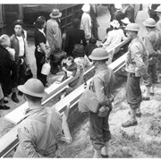 Helmeted soldiers stand guard as a group of Japanese who have just arrived on Pacific Electric Rail cars prepare to go to assigned quarters at the Santa Anita Assembly Center for the Japanese.  One young mother sits on a bench with her arm around her baby.