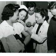Woman internee receives an inoculation. Three medical staff are administering it, while man in dark sweater is at the side.