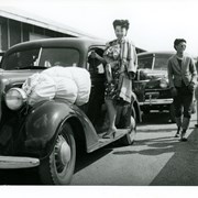 Japanese lady stands on running board of a car that apparently has just brought her and some of her family with their few allowed belongings to the Santa Anita Assembly Center for the Japanese.  Another car waits in line for the processing.  Two Japanese men and a policeman stand nearby.