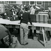In this photo, one staff member in dark sweater leans on large table set up to check belongings of new arrivals to Santa Anita Assembly Center for the Japanese.  Approximately twelve Japanese wait for the check.  Other staff members and police are standing about one large truck and two autos.