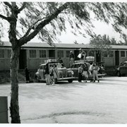Group of new arrivals at the Santa Anita Assembly Center for the Japanese prepare to unpack belongings into numbered quarters seen behind them.  Three cars are being unloaded.  About 16 Japanese can be seen in the vicinity. Pepper trees are seen in the area.