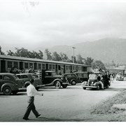 Seven cars and trucks are pulled up to housing units at the Santa Anita Assembly Center for the Japanese and the people are busily unpacking and preparing to move in. About 19 people can be seen in the photo.  The San Gabriel Mountains form a back drop.