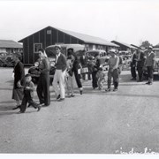 Thirteen newly arrived internees at Santa Anita Assembly Center for the Japanese, many of them small children. They are seen filing toward a processing point. Three staff members stand on right, identified by arm bands.  Automobiles are lined up behind. Housing units are also shown. The word INDUCTION is written across bottom of photo.