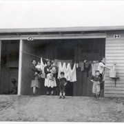 Three women, two holding babies, and two small children are seen standing in front of what appears to be a wash room area at Santa Anita Assembly Center for the Japanese.  Clothes hang on a line behind them.