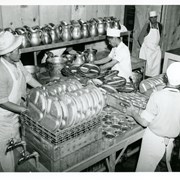 View of part of dishwashing operation at Santa Anita Assembly Center for the Japanese.  Photo shows 4 men working at stacking and readying metal plates and metal cups for washing.  Long shelf of large metal pitchers can be seen.