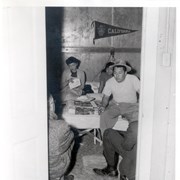 View of family group seen through the door in their living quarters at Santa Anita Assembly Center for the Japanese.  Lady reading the mail, young girl by bare light and man with hat sits on the end of the bed.  Two men sitting on low stools are by the door with backs to camera.