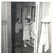 Two women stand inside a small room at Santa Anita Assembly Center for the Japanese; the one on the right is hanging clothes on a line, the other stands with her arms folded.  There is a parasol on the floor.