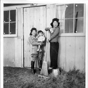 Two women standing outside one of housing units at Santa Anita Assembly Center for the Japanese.  Woman on the left is wearing a riding outfit and holding a small child.  The one on the right is standing on an overturned bucket and has a broom.