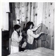 Young man at Santa Anita Assembly Center for the Japanese with broom at left, and young lady hanging up a sweater which she has just brushed.  There is a bed in the room also.