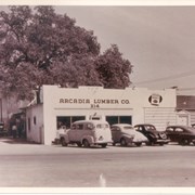 View from Santa Anita Ave toward east side at Arcadia Lumber Company at 214 N. Santa Anita Avenue.  Four cars and one pick-up truck are parked in front.  A large oak tree is behind office building.