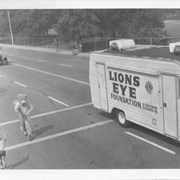 Lions Club van used in vision-screening program in Southern California drives in Diamond Jubilee Parade.  A lion-costumed person walking along side, reaches out to shake hands with a child.