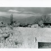 Small house and garage pictured on right side of photo.  The view is across field of grass toward San Gabriel Mountains in background.  Dramatic clouds fill the sky.  There is a bare, tall Lombardy Poplar pictured near the wash. Caption on photo says it is Santa Anita Wash, but viewer is not aware of it. It must lie along left side of photo.
