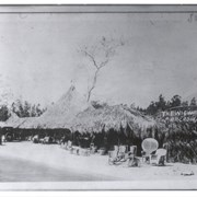 View toward west at front of a thatched-roof fruit and refreshment stand. On right is painted: THE WIGWAM, ARCADIA,CALIF. There are rattan chairs about and an automobile.  There is a sycamore tree seen behind the building. The eucalyptus trees of Santa Anita Avenue can be seen beyond.