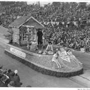 Close view of Arcadia's float during 1939 Pasadena Tournament of Roses Parade.  The designation in front reads FIRST SETTLERS 1839 and the name ARCADIA appears on side.  It has a small house at back of float with six people riding float.  None are identified.
