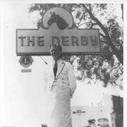 William O. Petersen, co-owner of The Derby standing beneath sign reading THE DERBY.  He is wearing white waiter's jacket and white apron.
