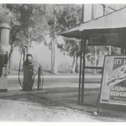 View toward west across Santa Anita Avenue near intersection with Lucile Street where a small service station was located (name of station and owner unknown).  Note large advertising poster on a stand promoting Gilmore Blu-Green Gasoline. Eucalyptus trees in center are prominent.