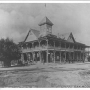 Oakwood Hotel.  On right edge of photo, Santa Fe steam engine is coming into view.  There are four men seen standing near Hotel.  There is a man in a buggy with two horses standing in shade of what appears to be an oak tree.  One lady stands on second floor porch.