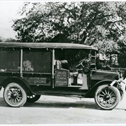 A REO truck with unknown driver standing in front of it.  Painted on side of truck: Santa Anita Rancho and Anoakia Breeding Farm, Anita M. Baldwin, Prop. Truck is under large oak tree.
