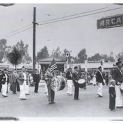 Members of Glenn Dyer Post of American Legion Drum & Bugle Corps are marching in Armistice Day Parade, 1934.  They are at intersection of First Avenue and Huntington Drive.  Arcadia Drive-In Market (aka Market Basket) can plainly be seen in background. None of marchers are identified.