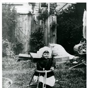 Baldwin M. Baldwin in child's metal seat outdoors.  He has on dark jacket and white bonnet. Wicker baby buggy shows on right of photo. Photo probably taken at Baldwin Ranch.